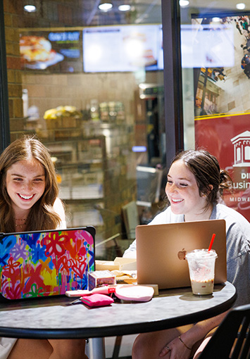 Two students sit together on their laptops, laughing, in the Dillard College of Business Administration lobby near Einstein Bagel.