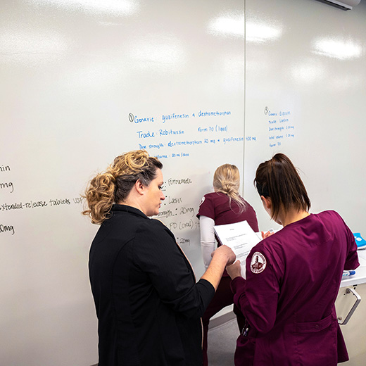 A nursing professor converses with one nursing student while another nursing student writes on the white board.