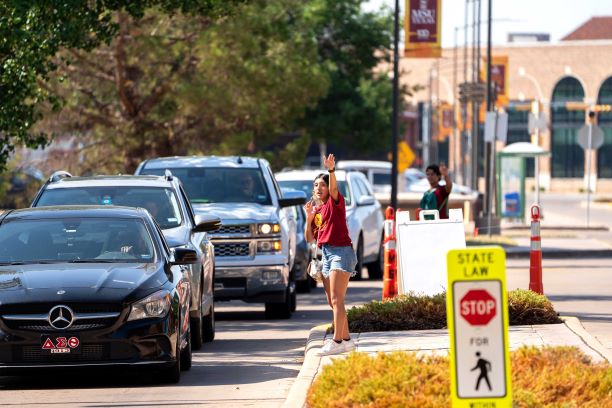 It is a sunny morning, as cars filled with students and their families line up and make their way down Louis J. Rodriguez Dr. on Move-In Day 2022. Two student volunteers in maroon Residence Life and Housing leadership shirts can be seen giving directions to the cars, as they make their way to the unloading zone in front of Legacy Hall and McCullough-Trigg Hall. MSU Texas banners with the Centennial Logo can be seen hanging from the light posts and the trees that line the street have dark green summer leaves. 