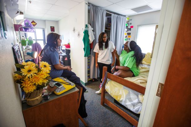 Killingsworth Hall Room interior view from the doorway looking into a double room. Three students are conversing in the 'A' side of the room. From left to right, one student is sitting on her desk which is situated on the left wall immediately seen as you come into the room. There are flowers, a lamp, and a variety of desk supplies on the desk. Behind her, light streams in from a window on the far side of the room. The small walkway between side 'A' and the window contains a metal shelf with shower supplies that hides the vanity sink from view and a micro-fridge on the opposite wall. Side 'B' cannot be seen from this angle but would run along the window wall mirroring side 'A'. The right side of the room branches off in an L shape. A second student stands in front of a large closet which is partially covered by a curtain hanging from a rod, but a variety of shelves and hanging clothes can be seen between the curtain gap. She is facing the third student who is sitting on a bed that takes up the space to the right along the same wall as the door. 