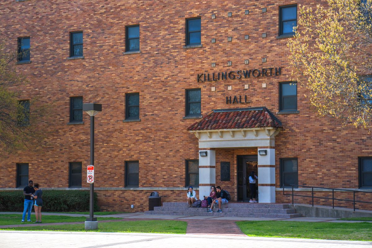 Students hanging out outside of Killingsworth Hall's east entrance exterior. The recessed entrance is flanked by white columns that contrast against the iconic MSU Texas red brick. Three steps lead up the entrance and a group of students can be seen sitting on these steps. Another student is swiping their card at the entrance to enter the building. A ramp leads from the entrance off-camera to the right. Two more students stand conversing on the right-hand side in the grass that separates Killingsworth from the brick Mustangs Walk. They stand under a tree that is just starting to shed its brown and yellow leaves for fall. 