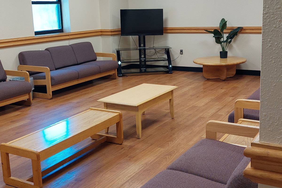 McCullough-Trigg Hall's first-floor lounge interior is seen from the doorway looking towards the far right-hand corner of the room. A television on a stand is in the far right corner near a window. Coaches, coffee tables, and a house plant line the walls creating a large space that can be rearranged for casual meetings, social functions, or relaxed study space.