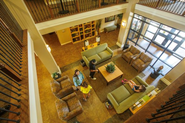 Sundance Court, looking down from the second story onto the main lounge, interior. Four students occupy the main lounge on the bottom floor of Sundance Court. The picture was taken from the second story lounge looking down. This square lounge area is situated in front of the glass double entrance to Sundance Court. Two cushioned chairs flanked by end tables sit against the windows on the edge of a square rug in the center of the tiled space. A coffee table is positioned on the rug with two green couches on either side and two more chairs mirroring the ones by the window creating a square seating area. Bookshelves line the far wall and the room is lit by various lamps, wall lights, and the afternoon sun coming in through the window. The first story's ceiling is open to the second story where another lounge can just be seen surrounding the first like a square balcony. Wood and metal railing line the balcony and a few high-top tables and chairs can be seen on the second story, but the rest of the second lounge is cut from view due to the angle of the shot.