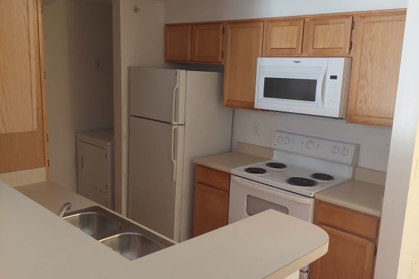 Sunwatcher Village, four-bedroom apartment kitchen, interior, move-in ready. The Sunwatcher Village kitchen is move-in ready meaning that the picture was taken prior to students moving in for the semester. The kitchen sits just off of the entryway. On the right, tan countertops and wooden cabinets line the wall. A stove/oven sits in the middle of the countertop with a microwave mounted above the stove connected to the ventilation system. A full-size refrigerator at the end of the kitchen area. A small wall divides the kitchen from the dryer. Metal shelving can be seen above the dryer. Opposite this wall is another counter space with an elevated bar area, but no wall, so the top section is open to the living room (not seen in this image). A two-compartment sink sits opposite the oven in the center of the countertop and a floor-to-ceiling cupboard sits across from the refrigerator. The washing machine (not in view) is across from the dryer next to the cupboard. 