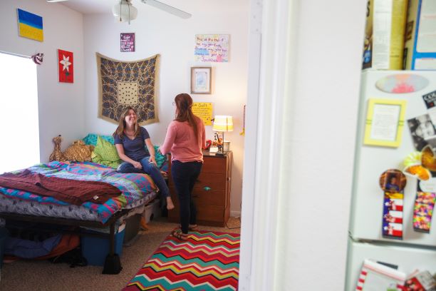 Sunwatcher Village, two-bedroom apartment, interior. Two students are talking in one of the bedrooms of Sunwatcher Village's two-bedroom/ two-bathroom apartments. The picture is taken just outside of the doorway of the bedroom and you can see part of the refrigerator on the right-hand side of the image. Through the doorway, you can see a full-size bed elevated off the ground allowing for storage underneath. It is positioned against the far corner of the room next to a large window. The dresser has been positioned next to the bed providing a space for a lamp and various items. There is a ceiling fan in the center of the room and a bright rug on the carpeted floor. The right side of the room is not seen due to the door being partially open.