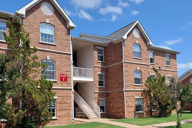 Sunwatcher Village's Building 2 exterior day. Building 2 is a three-story red-bricked building with a gray roof and white accents. The building is divided in two by a white metal staircase. Each floor has four windows on each side, and the blinds are closed so you cannot see inside. Large green bushes are on either side of a path leading through the grass to the covered stairwell. The blue sky is dotted with puffy white clouds.