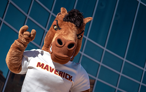 Maverick T Mustang holds up the Mustangs Hand Sign while posing outside of Centennial Hall on a bright, sunny day.