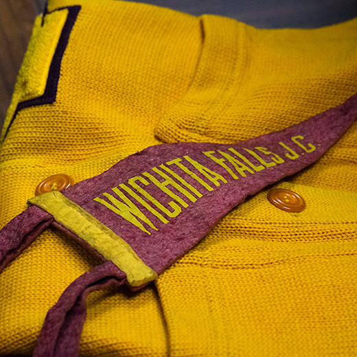 Image of a gold sweater with an embroidered maroon flag laid over top. Embroidered onto the flag is "Wichita Falls J.C." for when Midwestern State University was still named Wichita Falls Junior College.