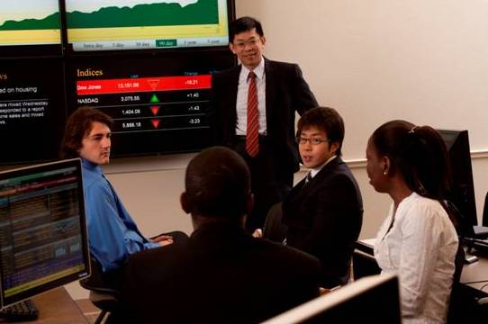 Faculty member and four students having a discussion near a NASDAQ stock monitor.