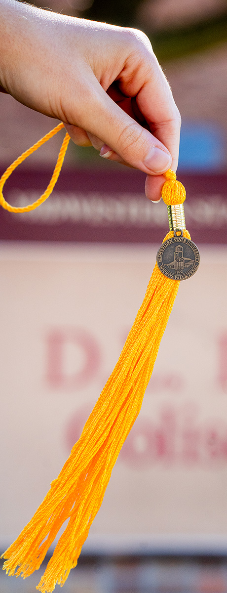 A student holds up a gold tassel outside of D.L. Ligon Coliseum, to represent commencement.