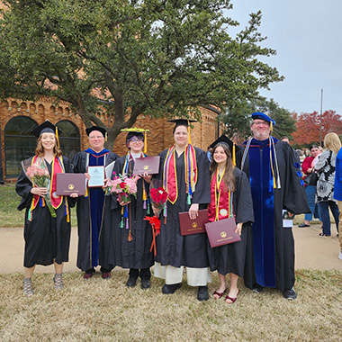 A group of four students happily pose with their professors after earning their degrees at commencement outside of D.L. Ligon Coliseum.