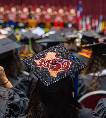 A student shows her decorated MSU Texas graduation cap as other graduates sit in front of and around her during MSU Texas commencement.