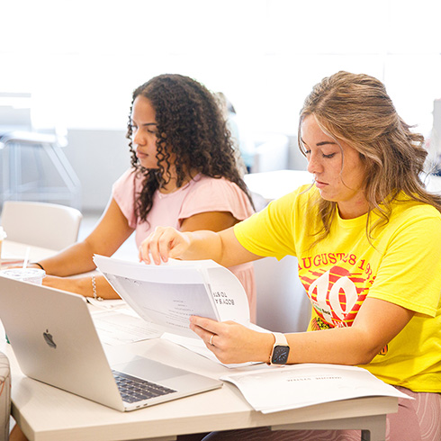 Two students work together with their laptops and paperwork.