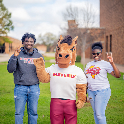 Two smiling MSU Texas students pose with Maverick T. Mustang in the center as they all hold up the Mustangs Hand Sign, outside, near Clark Student Center.