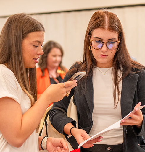 Two students converse over their phone and a printout during the career fair hosted by the Career Management Center, while employers engage with students in the background.