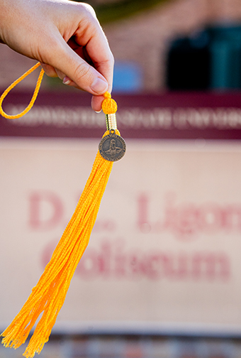 A student holds up a gold colored graduation cap tassel in front of the D.L. Ligon Coliseum sign.