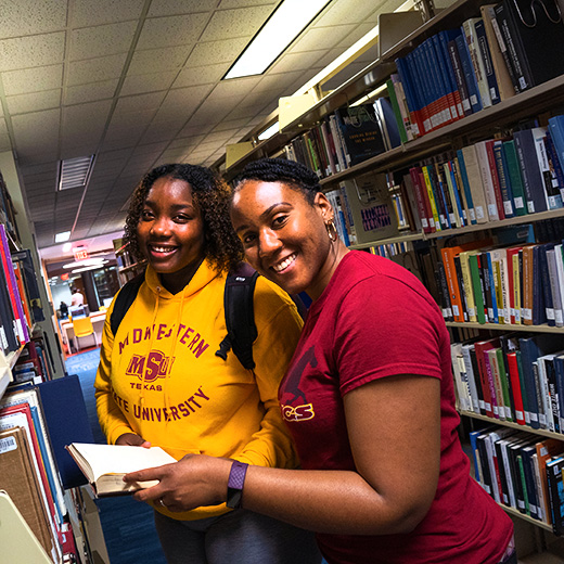 Two international students browse books together in Moffett Library, dressed in their Maroon and Gold t-shirt and hoodie, wearing bright smiles.