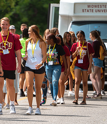 A group of students walks on campus on a bright, sunny day with the Midwestern State University shuttle in the background.