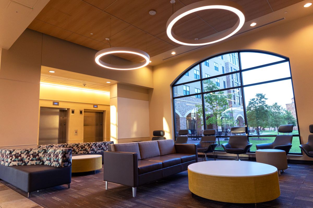 Legacy Hall's first-floor lounge interior. A large window looking out onto the Legacy Hall green space outside can be seen on the far right wall and the two elevators can be seen on the left wall. A variety of leather chairs, couches, circular ottomans, and coffee tables are spread out on the carpeted area. You could comfortably fit about 14 people in this seating area. Large circular light fixtures are hanging from the ceiling.