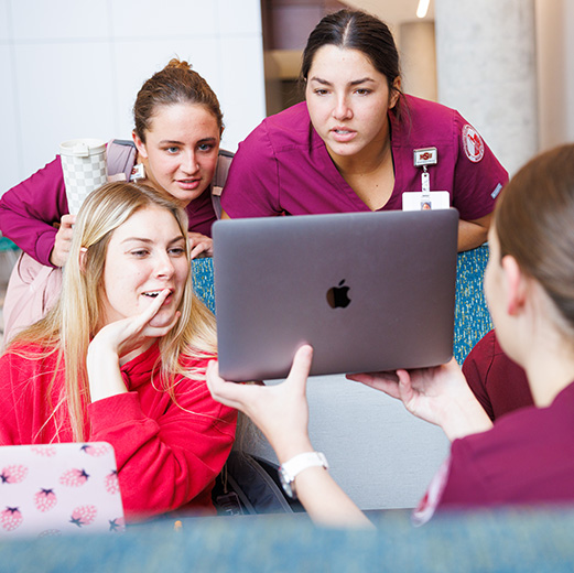 A group of four Health Sciences & Human Services students look at one student's laptop while gathered together at a table inside of Centennial Hall.