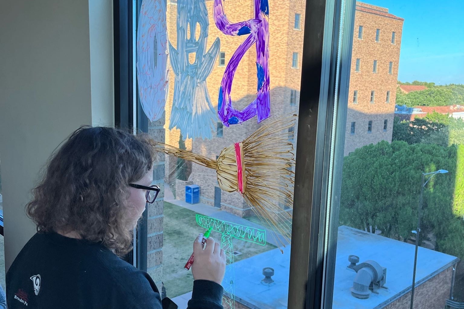 An honors student is decorating the Honors Lounge widows displaying a 'Boo' with a ghost as the middle 'O', a broom, and is working on the letter 'T'. Through the window, you can see part of Killingsworth Hall and the treetops around campus.