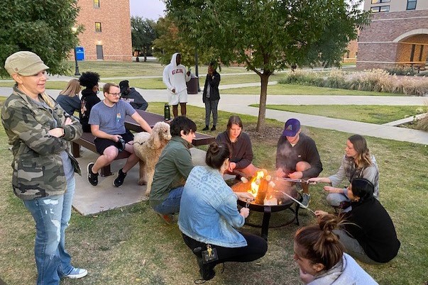 Several honors students are gathered outside in the Legacy Courtyard near a picnic bench. A fire pit has been brought outside into the courtyard and six of the students are roasting marshmallows around the fire. four more students are seated at the picnic table and several others are standing around the grass area talking. Paths crisscross through the grass of the courtyard behind the students. One of the wings of Legacy Hall can be seen in the background on the right and part of Killingsworth Hall can be seen on the left. The trees are still full of green leaves despite the cool afternoon temperatures that are causing many students to wear hoodies and jackets. Many of the lamp lights around the courtyard are just starting to come on as the sun sets off camera.