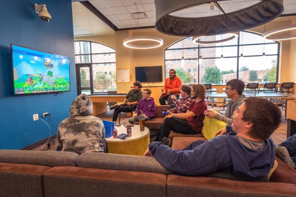A group of students from the Esports and Gaming Community is playing Super Smash Bros for the Nintendo Switch in the Legacy Commons on the first floor of Legacy Hall. The students are lounging on the large sectional couch and various chairs facing one of the TVs mounted in the commons. Under the TV are wall hook-ups for easy connection to their switch. A round ottoman is between the students and is covered in drinks and snacks. 