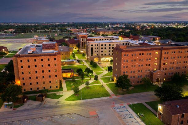 Legacy Courtyard as seen from a drone at dusk. The main walkway of Legacy Courtyard starts at the bottom left-hand corner and leads past Killingsworth Hall on the left and McCullough-Trigg Hall on the right. The main north entrance of Trigg can be seen along with all 6 floors. Just behind Trigg, Legacy Hall and Sundance Court can be seen as well as the Wichita Falls skyline stretching towards the horizon. The courtyard is lit with lamp posts along the walkways and the Legacy Commons located on the bottom floor of Legacy Hall, is brightly lit and the light spills out onto the main courtyard.