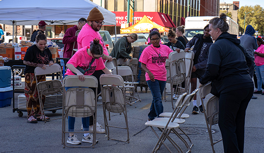 Students in hot pink Maverick's Day of Service t-shirts gather chairs on the sidewalk during an event in downtown Wichita Falls.