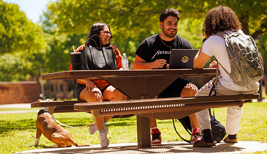 Three students happily converse near Clark Student Center, as their dog watches people walking by on campus.