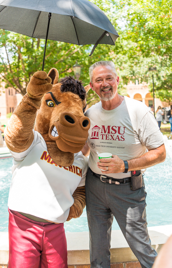 Maverick T. Mustang, the MSU Texas mascot, holds a black umbrella high above his head to shelter he and Interim President James Johnston from the sun. They gathered with member of the MSU Texas community near the Bolin Fountain located in The Quad to celebrate the University becoming the fifth members institution of the Texas Tech University System.